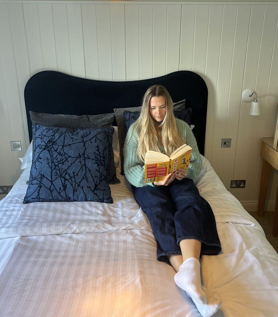 A blonde haired woman sitting on a bed with white duvet covers and navy pillows reading a book with a curved headboard behind her head.